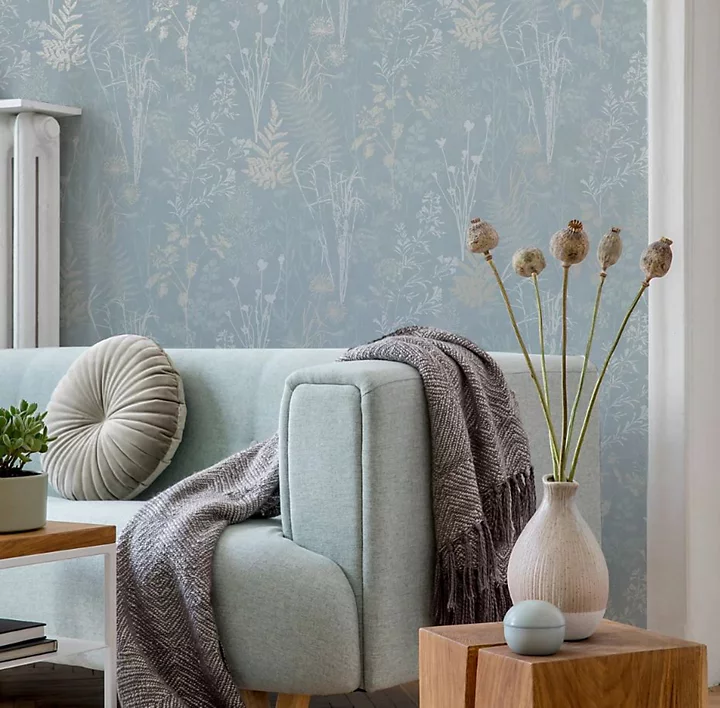 Giấy dán tường hình lá nhiệt đới màu xanh lá cây trong phòng khách với ghế sofa màu hồng nhạt và chăn màu xám nhạt.