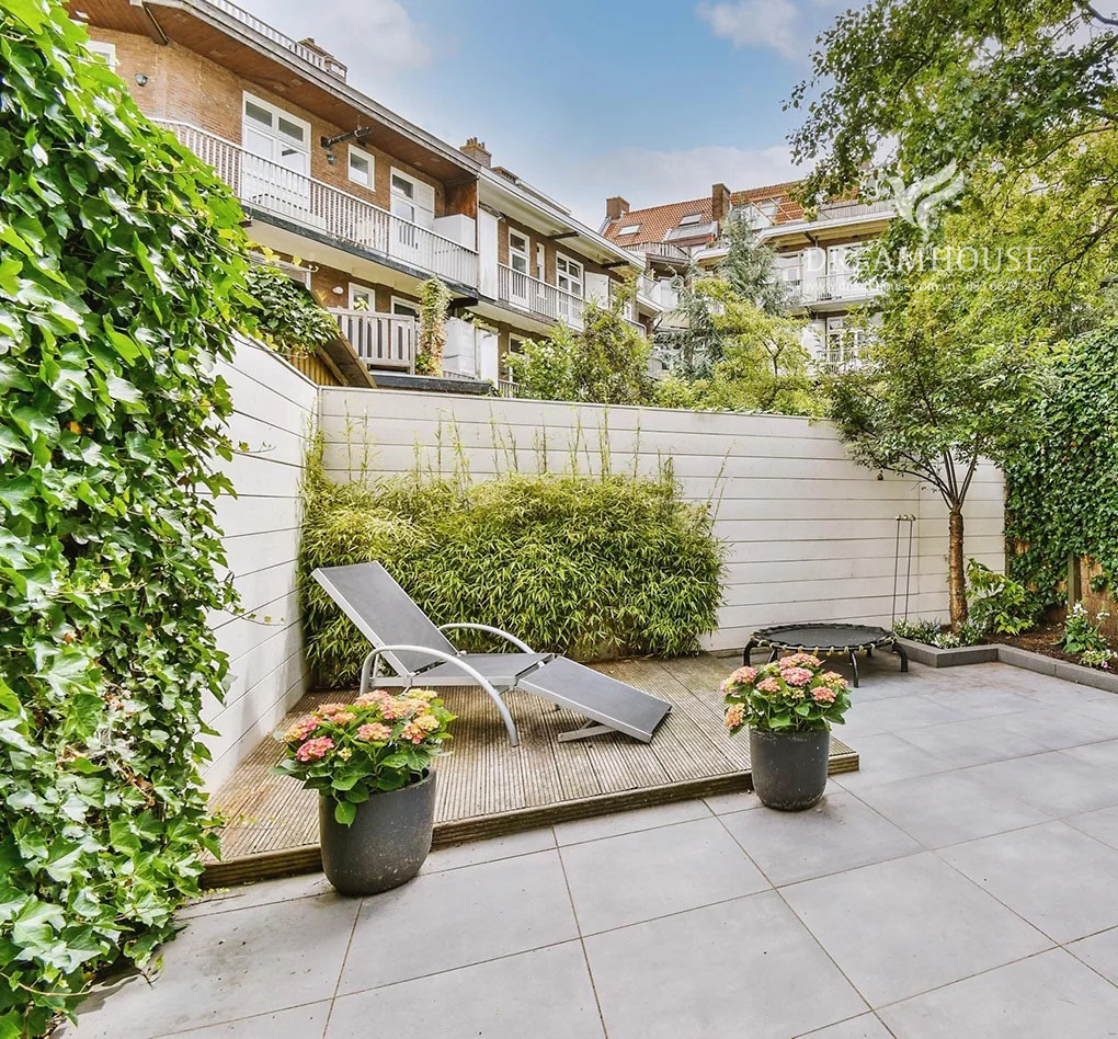 Khu vườn lát đá nhỏ có sàn nâng ở góc, có ghế tắm nắng và cây trồng trong chậu.